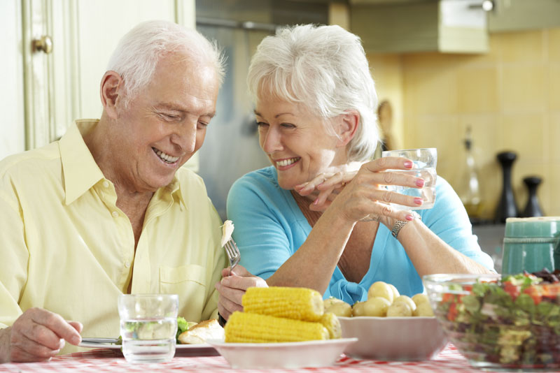 a couple smiling at each other while they are eating their dinner because their implant supported dentures are giving them a more stable smile.